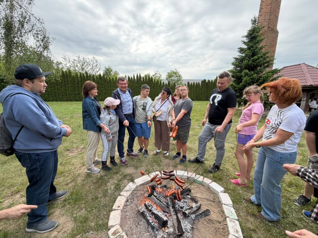 Grupa uczestników i opiekunów stojących wokół ogniska i piekących kiełbaski podczas spotkania integracyjnego