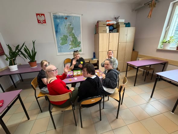 Grupa młodych ludzi oraz ich opiekunka siedzą przy stoliku z rozłożoną grą planszową i uśmiechają się w stronę aparatu, a dwie osoby machają ręką. Nad nimi na ścianie wisi godło Polski oraz mapa Wielkiej Brytanii.