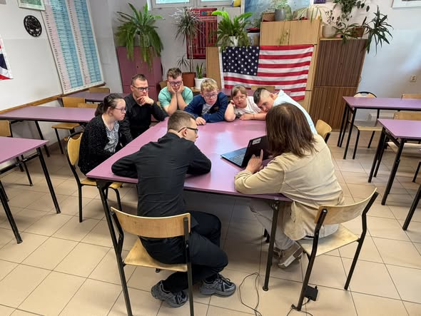 Prowadząca zajęcia kobieta w beżowej marynarce pokazuje coś na ekranie otwartego laptopa grupie młodzieży siedzącej przy dużym, złączonym stoliku. Uczestnicy z uwagą obserwują ekran. W tle na szafie wisi flaga Stanów Zjednoczonych