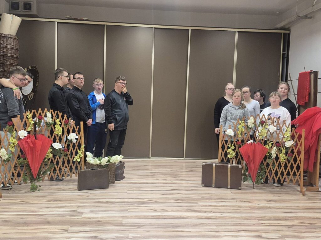 Grupa podopiecznych stojących w sali przygotowanej do zajęć teatralnych, pomiędzy dekoracjami z kwiatami i czerwonymi parasolami