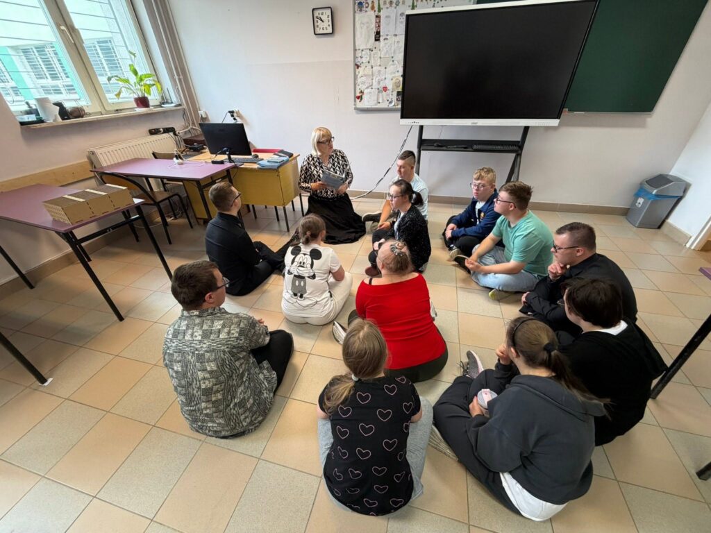 Grupa kilkunastu uczestników siedzi w kręgu bezpośrednio na kafelkowej podłodze sali lekcyjnej. Przed nimi stoi nauczycielka w ciemnej spódnicy i wzorzystej bluzce, trzymając w rękach książkę lub materiały edukacyjne i rozmawiając z grupą.