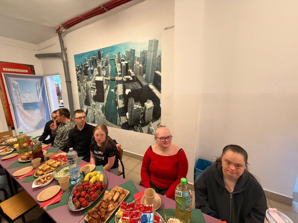 Bliższe ujęcie na uczestników siedzących przy stole z poczęstunkiem w korytarzu. Na pierwszym planie widać dziewczynę w ciemnej bluzie oraz jej koleżankę w czerwonej bluzce i okularach. Na stole królują półmiski pełne świeżych truskawek i ciast.