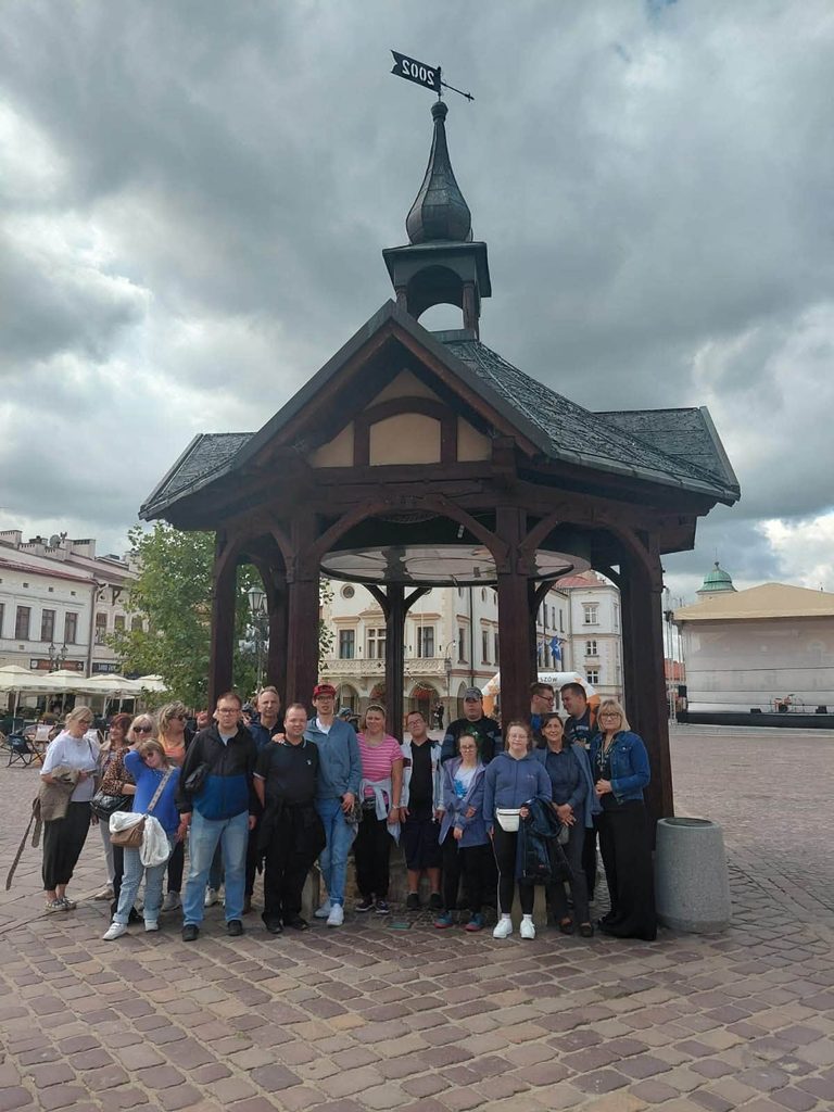 rupa uczestników wycieczki pozuje do wspólnego zdjęcia pod drewnianą studnią znajdującą się na rynku w Rzeszowie. W tle widoczne są zabytkowe kamienice oraz płyta rynku