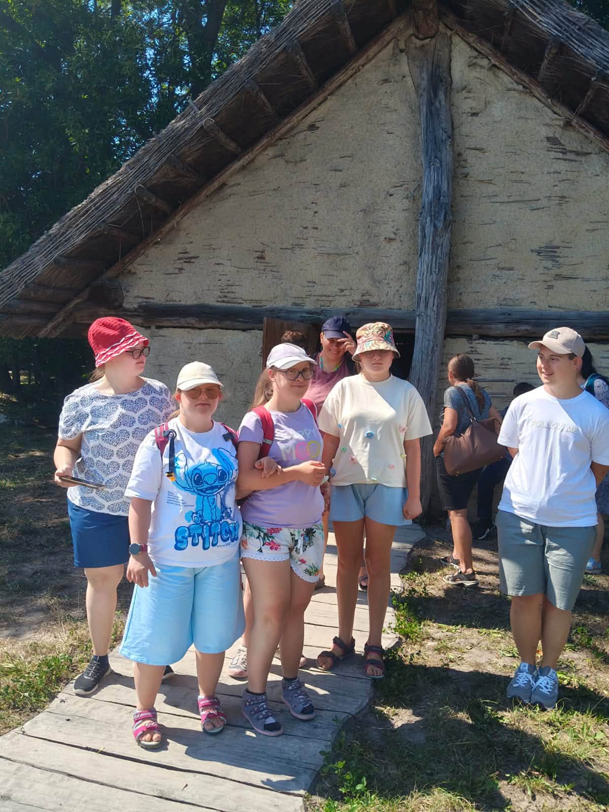 Grupa uczestników zwiedza rekonstrukcję osady Gotów w Masłomęczu, stojąc przed tradycyjną chatą.