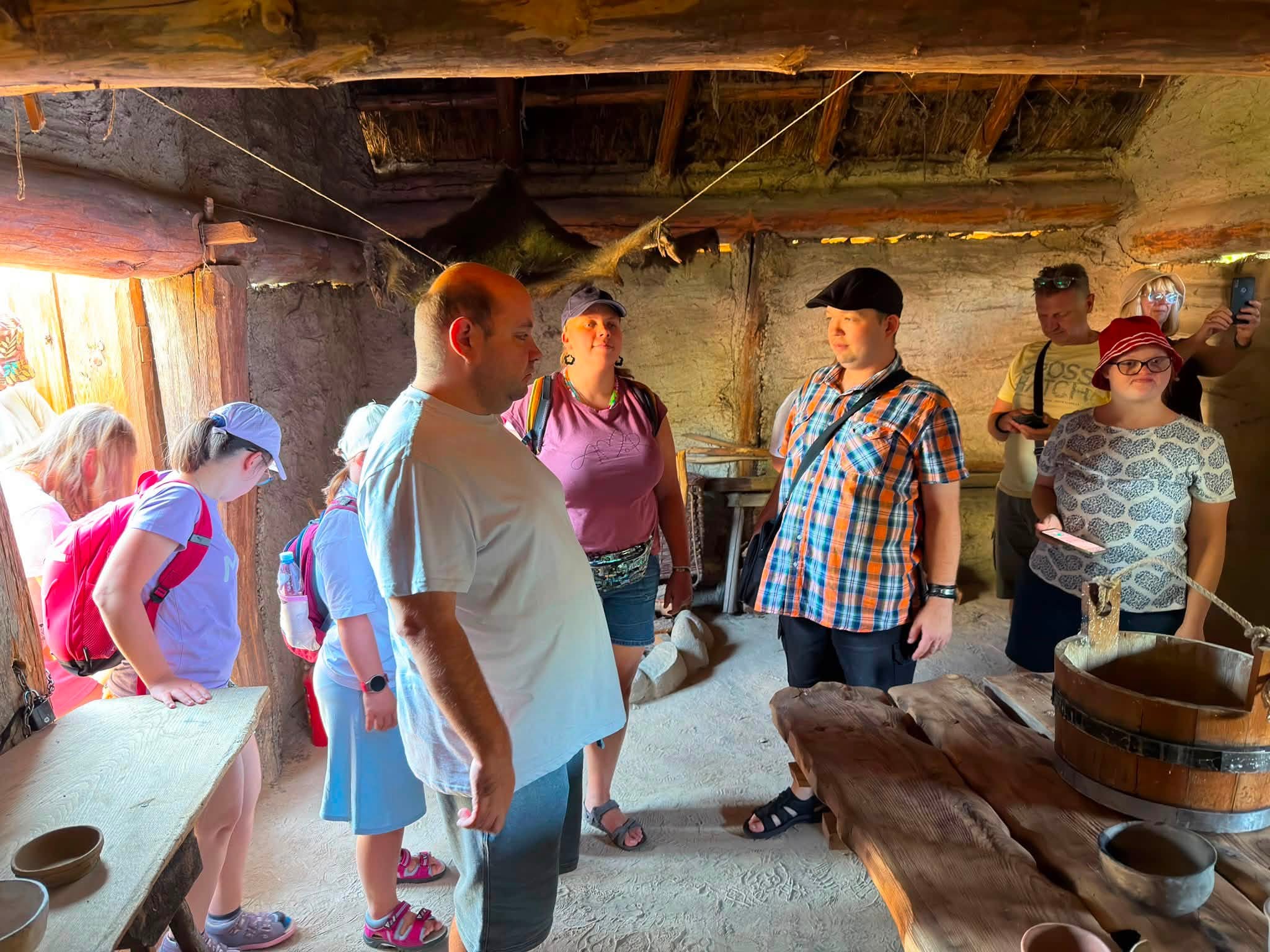 Grupa zwiedzających słucha opowieści przewodnika we wnętrzu zrekonstruowanej chaty w osadzie Gotów