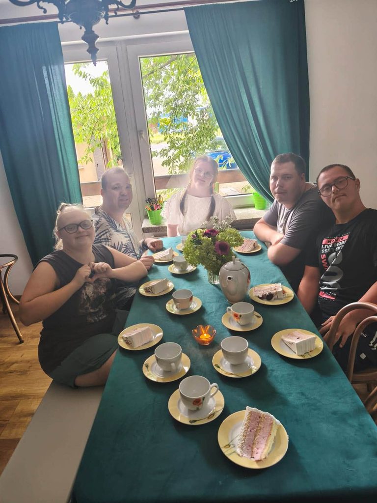 Uczestnicy świetlicy siedzą przy wspólnie nakrytym stole podczas spotkania urodzinowego. Na stole znajdują się filiżanki, talerzyki z kawałkami tortu oraz dzbanek do napojów. W tle widoczne jest okno z zasłonam