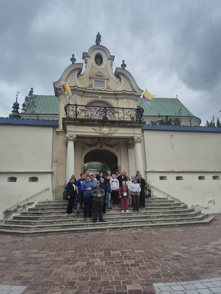 Grupa uczestników wycieczki stoi na schodach przed wejściem do Sanktuarium w Leżajsku. W tle widoczna jest zabytkowa brama i fragment kompleksu klasztornego.