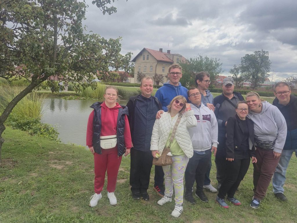 Uczestnicy wycieczki pozują do wspólnego zdjęcia nad stawem. W tle znajdują się drzewa, zabudowania oraz zachmurzone niebo.