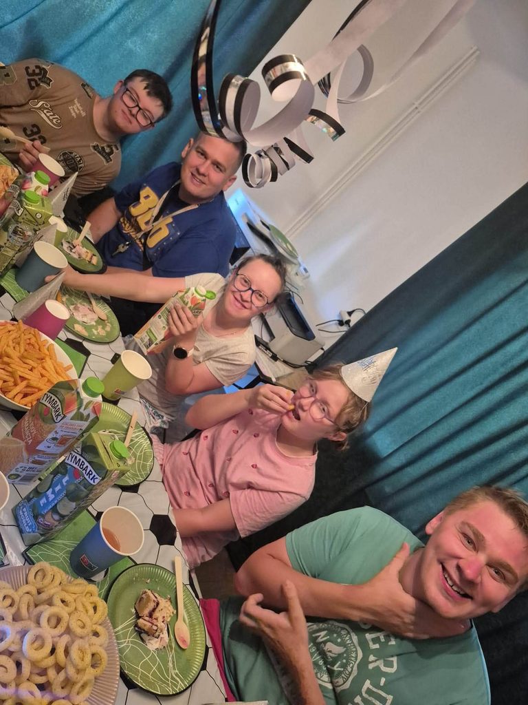 Uczestnicy spotkania urodzinowego siedzą przy wspólnie przygotowanym stole. Na stole znajdują się przekąski, kawałki ciasta, soki, jednorazowe naczynia oraz dekoracje urodzinowe. W pomieszczeniu widoczne są ozdobne serpentyny zawieszone pod sufitem