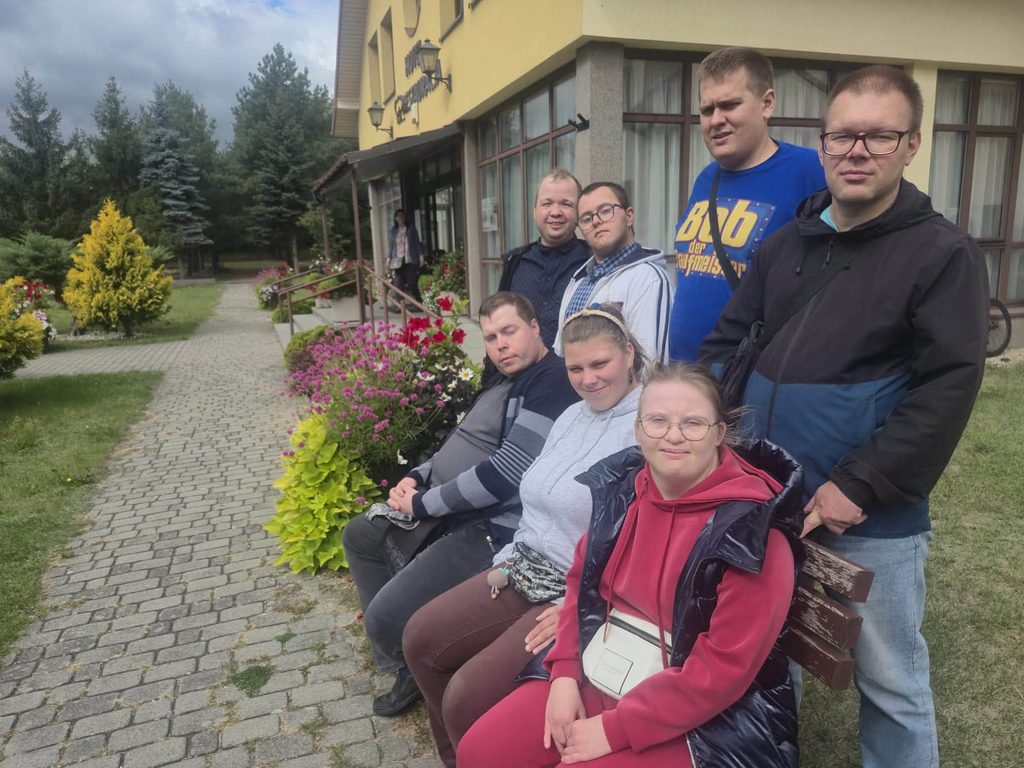 Grupa uczestników odpoczywa przed budynkiem ośrodka. Wokół widoczne są kwiaty, alejka oraz teren zielony.
