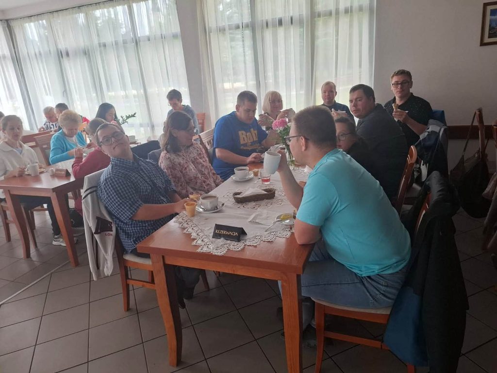 Uczestnicy wycieczki siedzą przy wspólnym stole podczas obiadu w restauracji. Na stołach znajdują się napoje i zastawa stołowa.